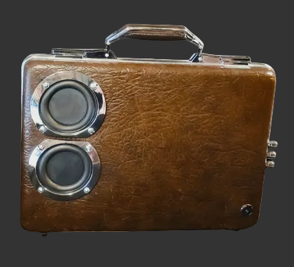 A vintage-style brown leather briefcase retrofitted to speaker enclosure with a top handle, two circular speaker grilles with chrome bezels, and a small metal latch on the right side, shown against a plain white background.